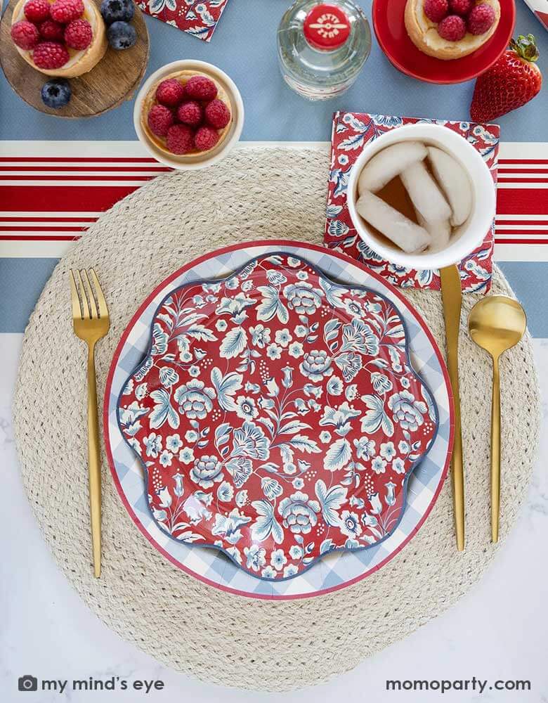 An elegant Fourth of July party table features Momopartys's Hamptons Collection by My Mind's Eye including 10" Chambray Gingham Paper Plate, Hampton Floral Wave Plate and napkin on a round white placemat. In the middle of the table there are blue and red treats and drinks on the Hampton striped paper runner, making this a perfect party inspo for a 4th of July party or Memorial Day celebration.