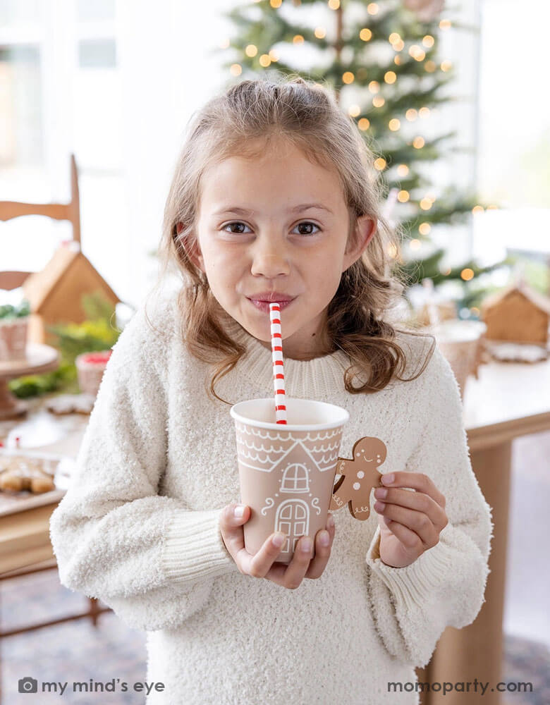 A cute girl in a white sweater holding drinking hot chocolate from Momopartys's 12 oz Gingerbread Handled Paper Cup with a festive gingerbread man handle. She's sipping her drinks with a red and white striped reusable straw. In her back there's a table filled with gingerbread houses and a Christmas tree with lights in background, which creates a cozy yet festive vibe for a whimsical Christmas party for kids this Holiday season.