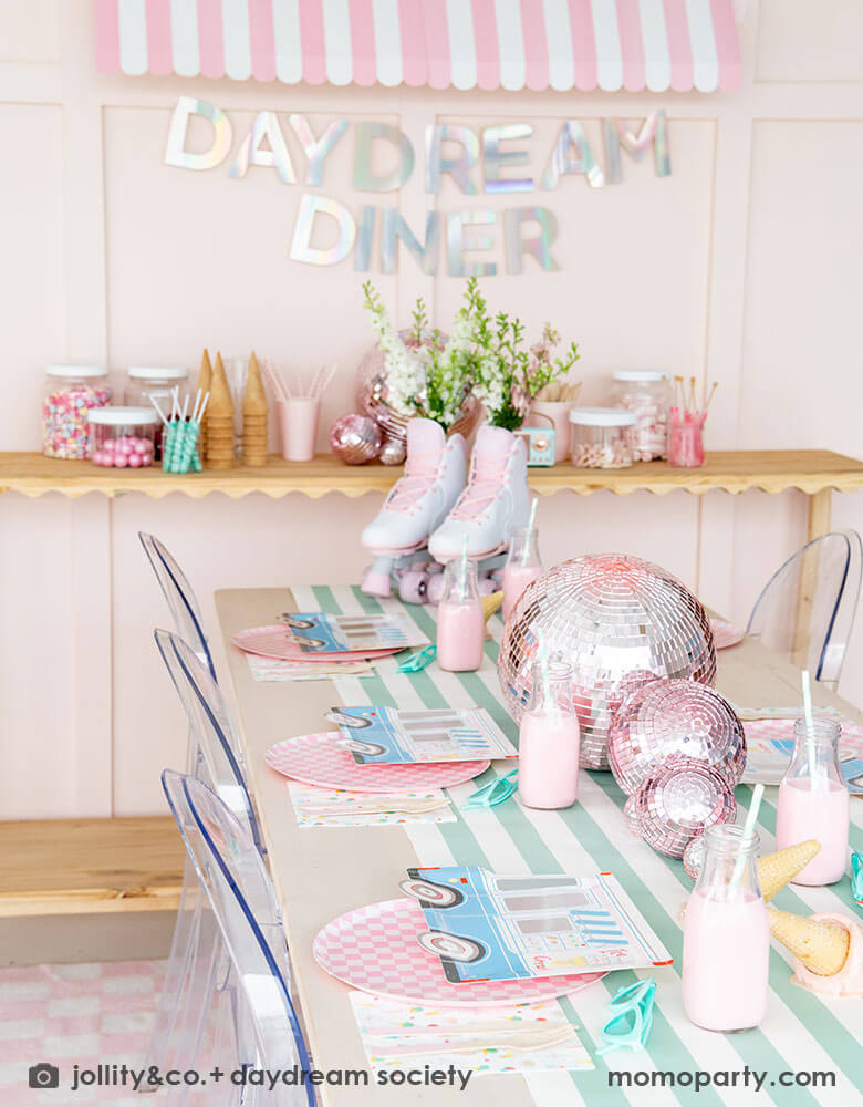 An adorable kid's ice cream themed birthday party tablescape featuring Momopartys's ice cream truck shaped plates, the pink checkered plates, the ice cream icon napkins on a mint striped table runner. On the table there's a roller skate and pink disc balls as centerpiece. Behind the table there's a sundae station with sprinkle toppings, ice cream cones, adorned with a retro pink striped awning as a small ice cream shop, making this a cute and festive ice cream party set up for kids birthday celebration.