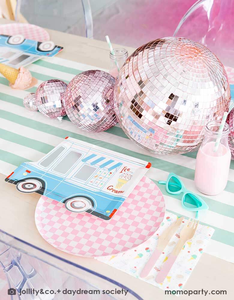 An adorable kid's ice cream themed birthday party tablescape featuring Momopartys's ice cream truck shaped plate, the pink checkered plate, the ice cream icon napkin and pink wooden cutlery on a mint striped table runner. Around the placeset there are some summer inspired decoration like a pair of mint sunglasses, pink disco balls, a pair of pink roller skates, some milk bottles with gum ball pink drinks, and some ice cream cone decorations, making this a cute, fun and festive ice cream social.
