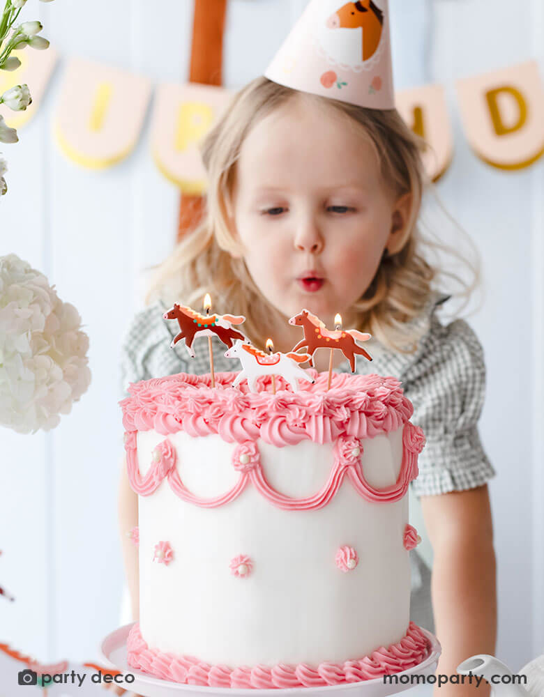 A preschool aged girl in her green gingham dress, wearing Momopartys's floral pony birthday party hat and blowing Momopartys's floral pony candles by Party Deco on a pink and white vintage buttercream cake. Behind her is a blush happy birthday banner hung on the wall. An adorable pony horse themed birthday party set up for a little girl.