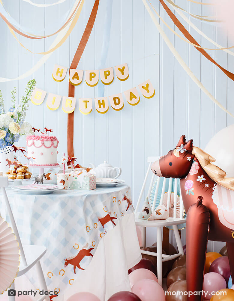 A pony themed birthday party table featuring Momopartys's floral pony party supplies including party cups, plates, cupcake toppers, napkins treat boxes and paper straws. In the middle of the table is a pink and white buttercream cake topped with pony shaped birthday candles, along with the blush happy birthday party banner hung on the wall. Next to the table stands an adorable pony shaped foil balloon adorned with floral designs. Making this a perfect inspo for girl's horse themed birthday celebration.