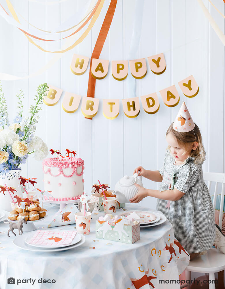 A preschool girl with a birthday party hat in a gingham dress pouring tea from a tea pot on a party table featuring Momopartys's floral pony themed party supplies including floral pony party cups, plates, cupcake toppers, napkins treat boxes and paper straws. In the middle of the table is a pink and white buttercream cake topped with pony shaped birthday candles, along with the blush happy birthday party banner hung on the wall - making this a perfect info for a girl's horse themed birthday party.