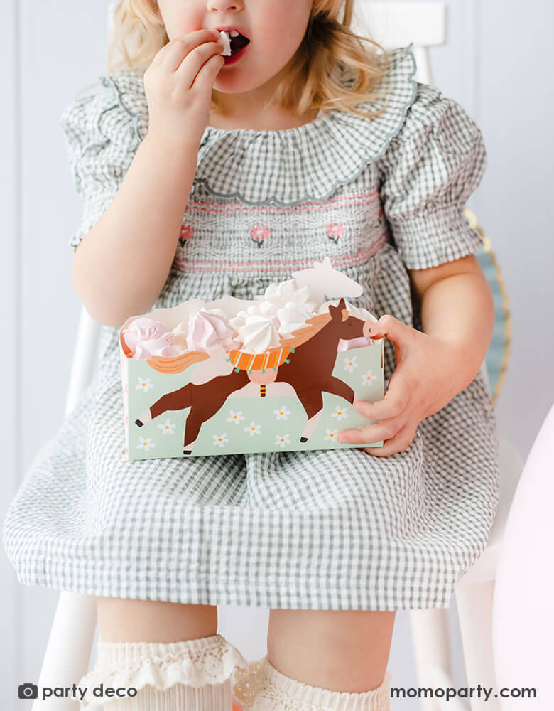 A preschool aged girl in a green gingham checkered dress sitting on a chair holding Momopartys's floral pony treat box by Party Deco. In the snack box are pink and white meringue - a great party snack idea for girl's horse themed birthday party.