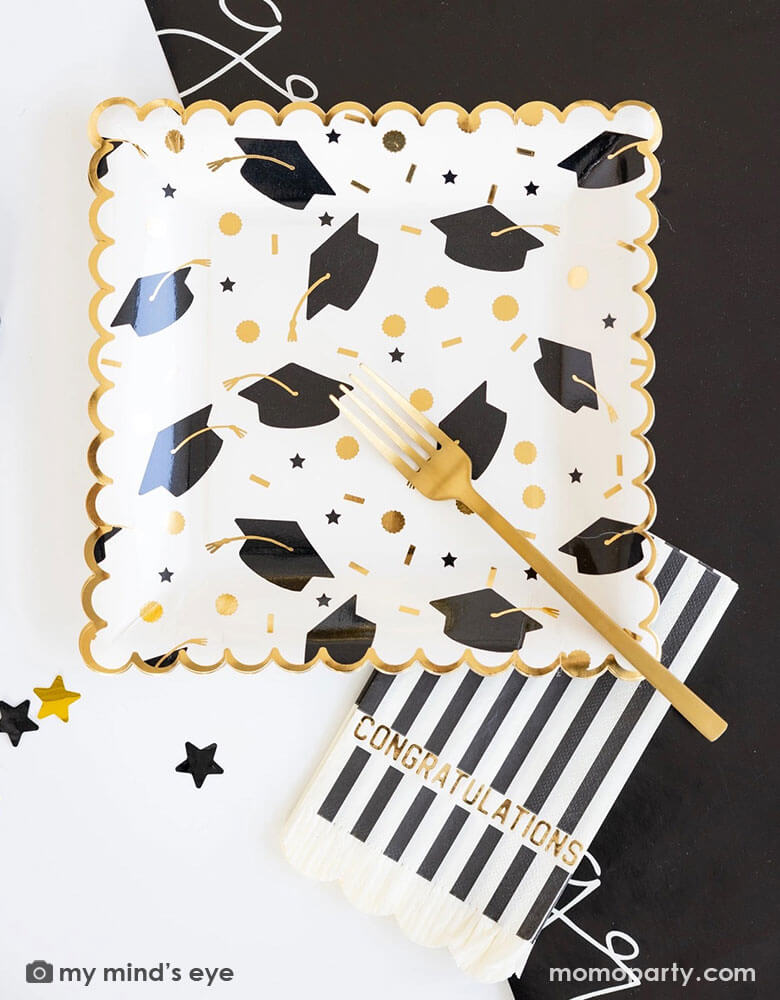 A graduation party table features Momopartys's 9" x 9" black and gold cap scatter paper plates, congratulations black and white striped dinner napkins, and congratulation black paper runner along with some black and gold star shaped confetti by My Mind's Eye. Making this a great inspo for a modern and stylish graduation party tablescape.