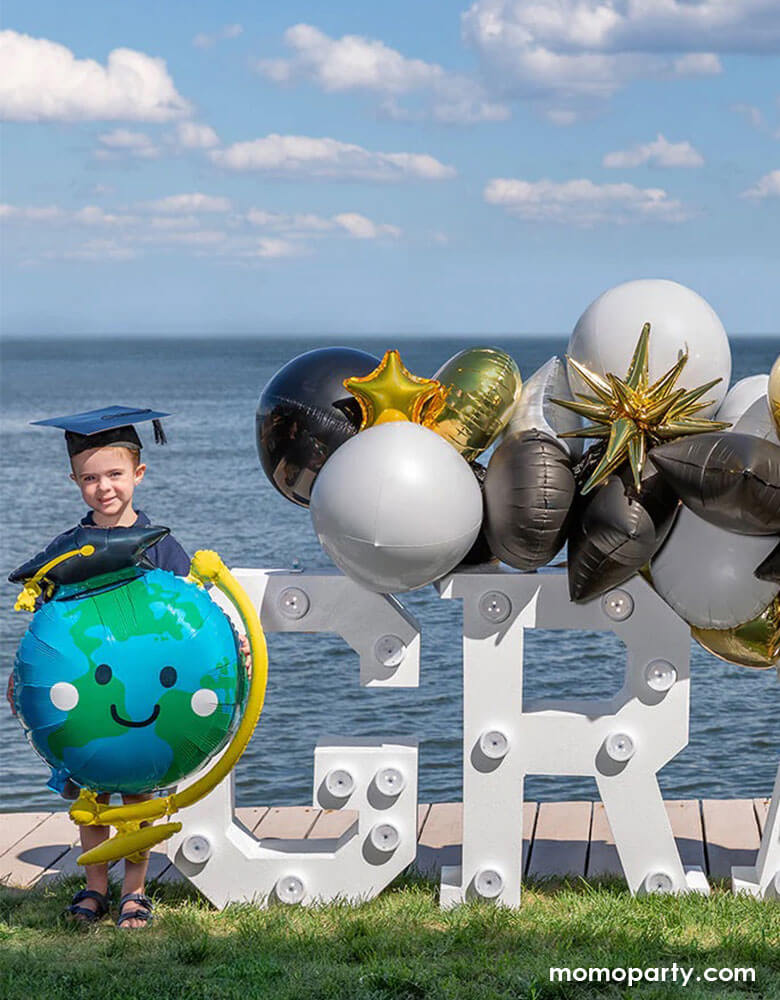 A preschool or kindergarten age kid holding Momopartys's 29" Graduation Fun Globe Shaped Foil Balloon standing next to a graduation marquee sign decorated with graduation themed balloon garlands in black, gold and white to celebrate his graduation day.