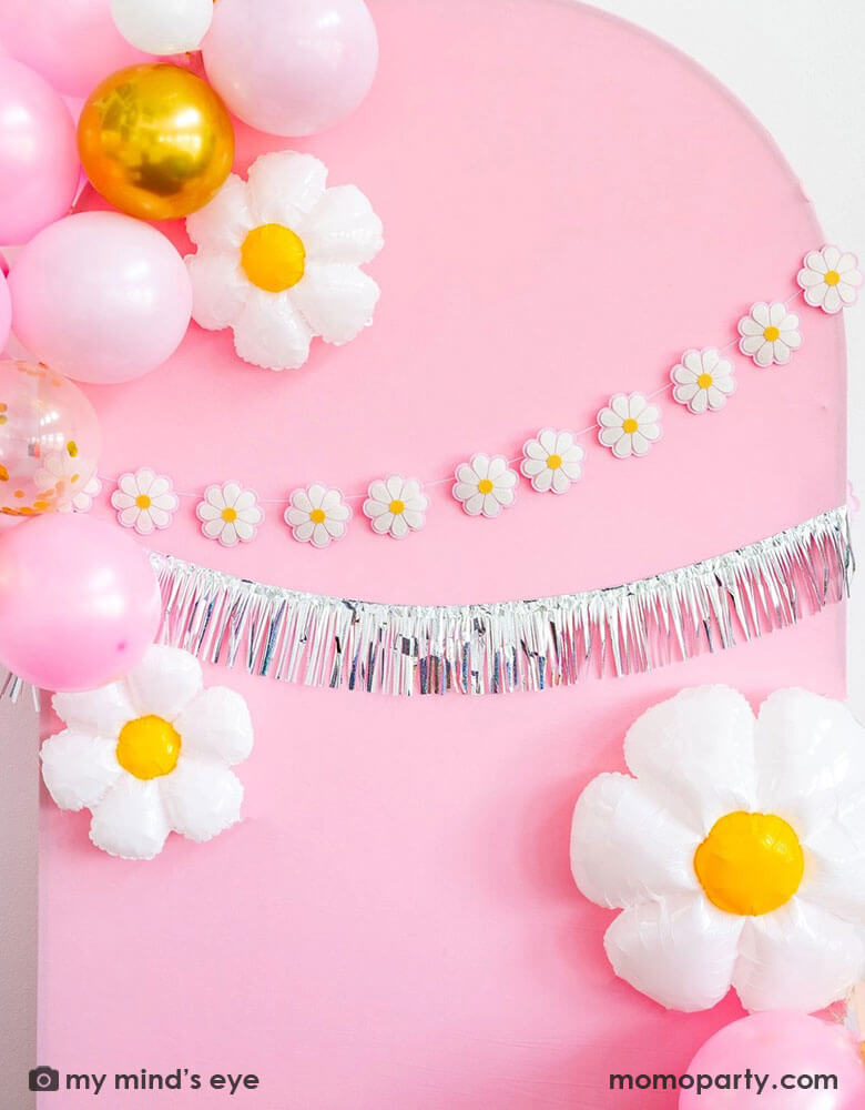 A pink wooden party backdrop wall decorated with Momopartys's daisy felt banner and silver fringe banner set. Around it are a organic balloon garland in pink, white and gold colors, with some daisy shaped foil balloons in different sizes, making this a perfect inspo for a festive and adorable decoration for kid's daisy themed birthday party or a retro inspired bash!