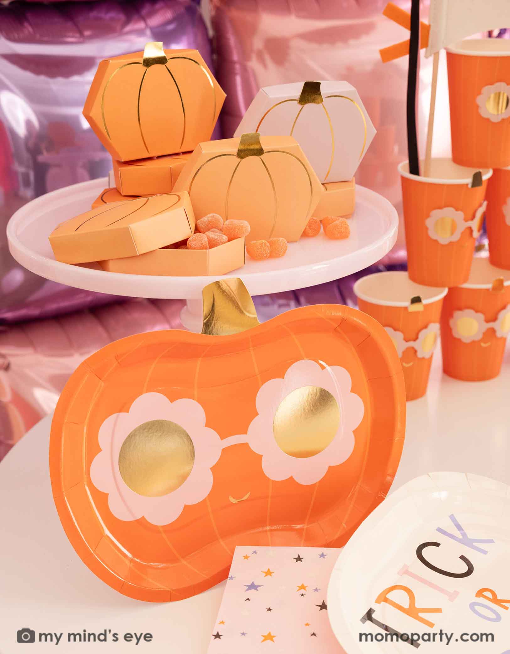 A Halloween party table featuring various Halloween-themed party supplies from Momopartys including pumpkin-shaped plates, party cups, trick or treat plates, napkins and some pumpkin shaped treat boxes filled with orange gummies on a pink cake stand, in front of a pink and lilac balloon wall background.
