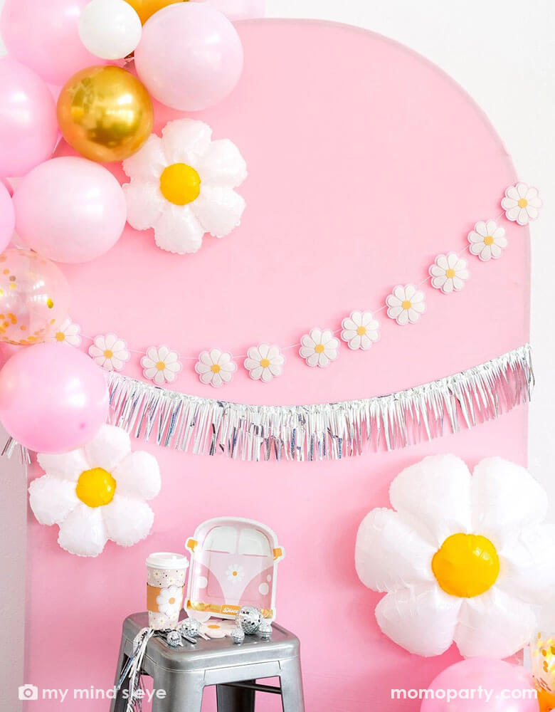 A pink retro daisy themed party decoration set up featuring Momopartys's disco daisy themed party supplies including VW van shaped plates, party cups, daisy shaped napkins on the stool. In the back there's a pink curve wooden backdrop adorned with My Mind's Eye's 6ft Disco Daisy Puffy Felt Banner and a silver fringe garland. Along the backdrop there are also a balloon garland with gold, pink and white balloons and daisy shaped foil balloon. Making this a great inspo for a groovy daisy hippie themed bash!
