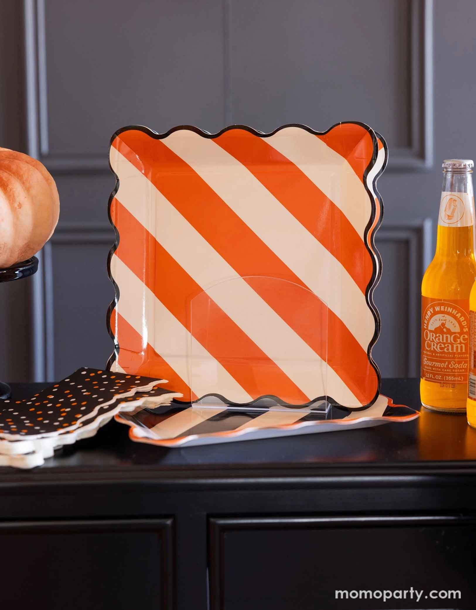 A black tabletop featuring Momopartys's Orange Striped Plate with orange and white diagonal stripes with wavy black edge. Around the plate there are some Halloween-themed items including orange soda bottles, pumpkins, black star confetti napkins.