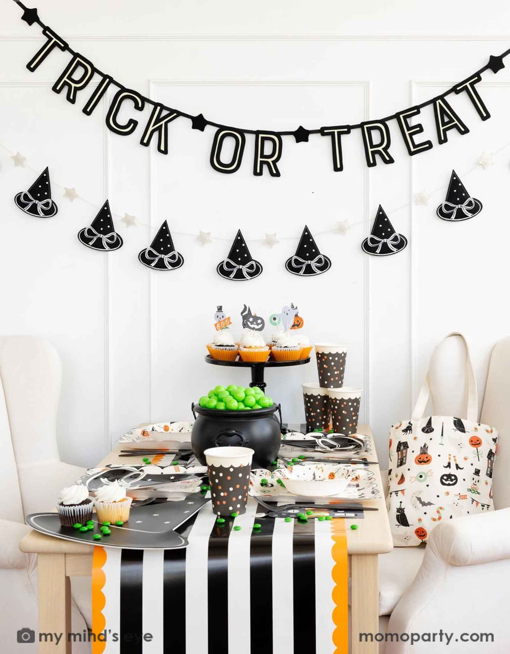 A Halloween-themed party with Momopartys's 6 ft 'Trick or Treat' glow in the dark banner and the witch's hat garland hung on the white wall. There's a table decorated with multiple Halloween party supplies by Momopartys including witch's hat shaped bamboo tray, party cups and orange, black and white striped table runner draping from the table which some orange and black balloons next to. A cute and simple set up for kids to celebration Halloween at home.