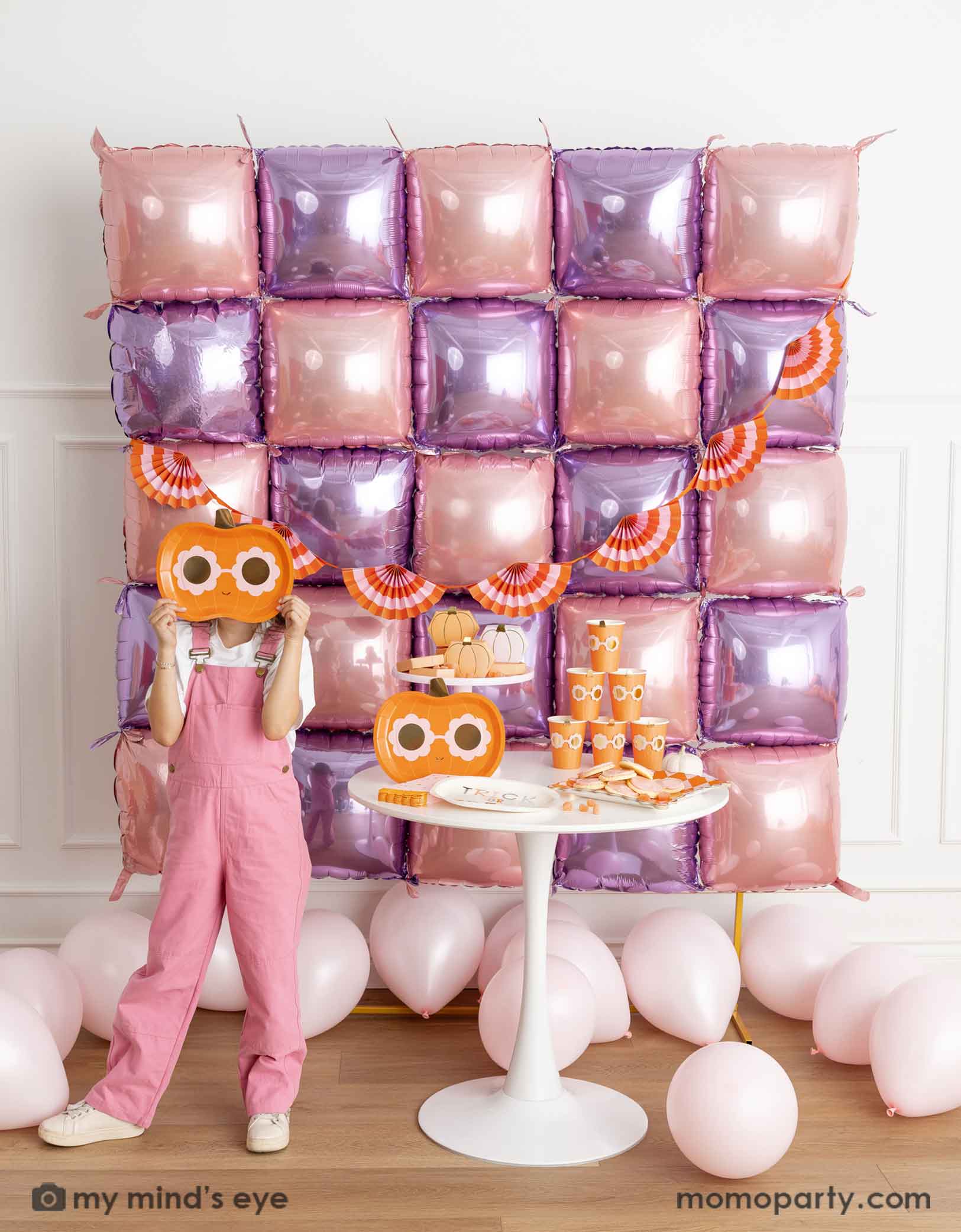 A girl in her pink overalls holding Momopartys's pumpkin-shaped plate standing in front of a decorative wall with pink and purple foil balloons adorned with an orange and pink bunting garland. Next to her is a white round table displaying groovy halloween pumpkin party tablewares including party plates, cups, napkins and more. On the floor are some pale pink latex balloon, making this a festive kid's friendly pink Halloween celebration.