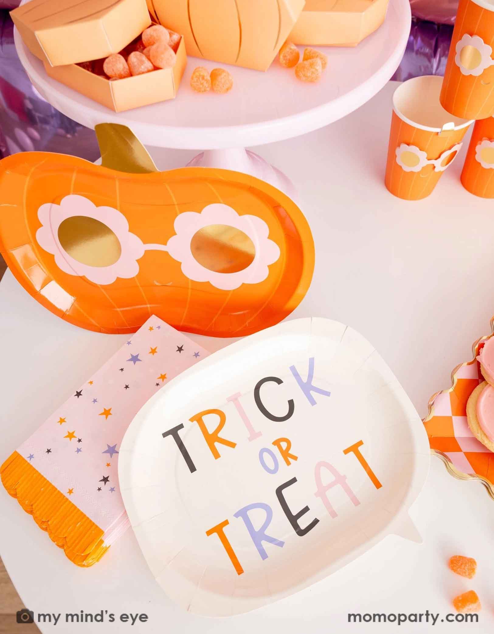 A Halloween-themed table featuring various pink Halloween themed tableware from Momopartys including a 'Trick or Treat' plate, the groovy pumpkin-shaped plate and party cups, plus pink star with orange fringed napkins. Making this an adorable tablescape for a cute Halloween party celebration.