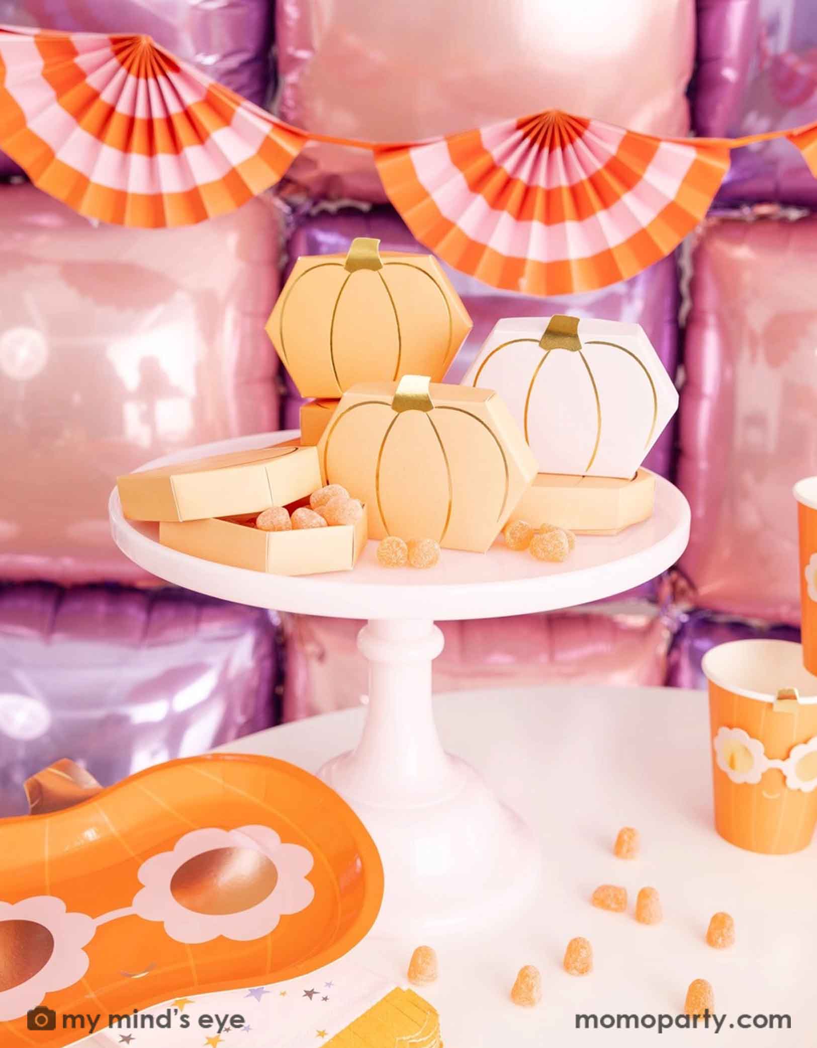 A festive Halloween-themed party setup with Momopartys's Pumpkin-shaped boxes by My Mind's Eye filled with orange gummies on a pale pink cake stand with festive pink Halloween decorative background with foil balloon wall and bunting garland. On the table there are groovy pumpkin shaped plates and party cups and some candy corn spread-out. A sweet and kid's friendly set up for the spooky season.