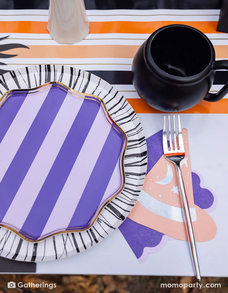 A close up shot of a festive Halloween witch themed party table featuring Momopartys's Bewitching purple side plates on a black and white striped dinner plates, bewitching witch shaped hat napkin and a cauldron shaped cup on a black and orange striped table runner. A perfect inspo for Halloween witch themed table for this spooky season!