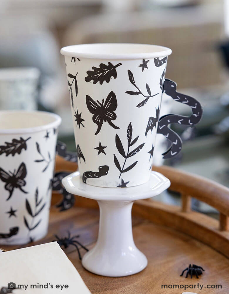 A close up shot of Momopartys's 12 oz Snake Handle Party Cups by My Mind's Eye on a wooden table covered with spider decoration and confetti. These spooky cups not only come in a fun snake handle design, but they will also make your party stand out in a unique way. Don't settle for boring party cups, add a touch of fun with these cups!