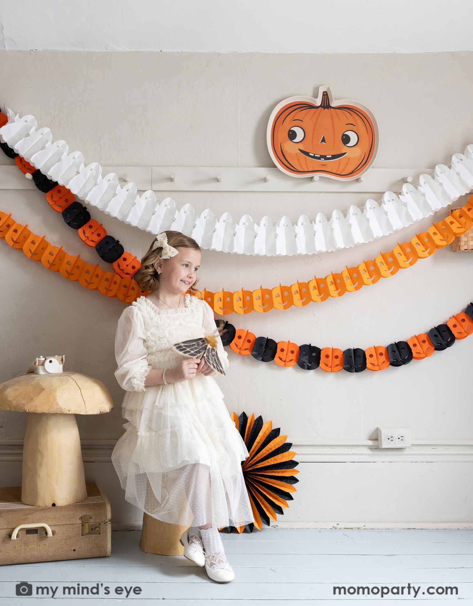 A girl in a vintage inspired white tulle dress sitting next to a white wall with coat hanging rack decorated with Halloween-themed decorations including Momopartys's pumpkin-shaped bamboo tray, ghost tissue garland, orange Jack-o'-lantern tissue garland, plus an orange and black Jack-o'-lantern tissue garland hung on the wall. Underneath is a cat figurine on a bunch of wooden mushroom stools which are next to a orange and black tissue paper fan decoration.