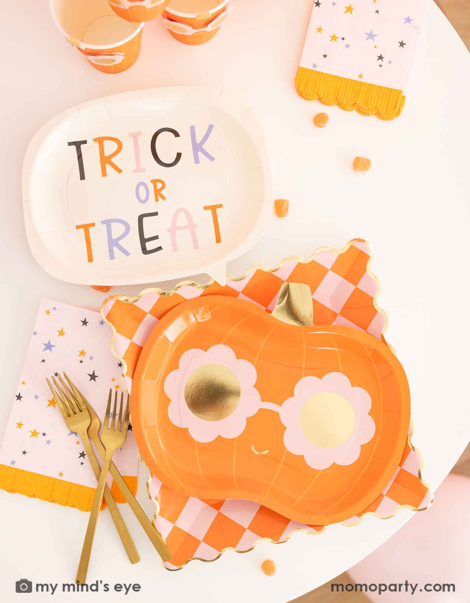 A Halloween-themed table featuring various pink Halloween themed tableware from Momopartys including a 'Trick or Treat' plate, a orange checkered dinner plate, a groovy pumpkin-shaped plate and party cups, plus pink star with orange fringed napkins. Making this an adorable tablescape for a cute Halloween party celebration.