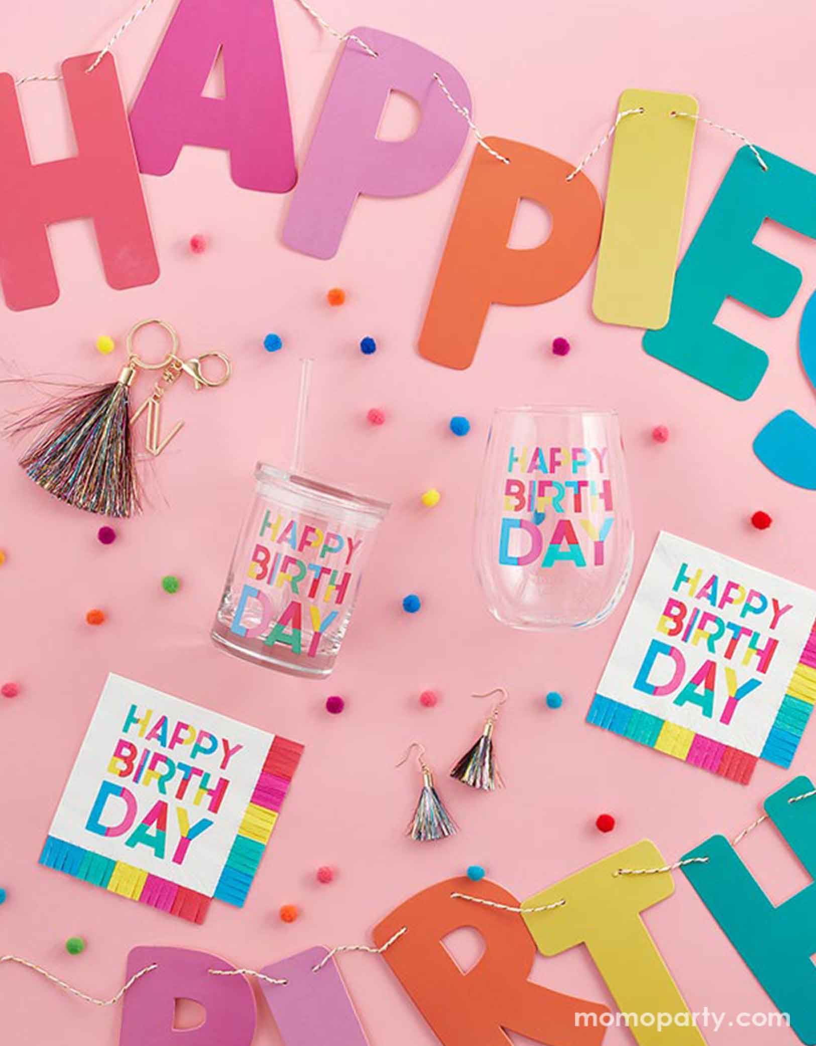 A overhead shot of a festive birthday party table featuring colorful birthday party supplies including rainbow colored fringed napkins, "Happy birthday" drinking cups and Momopartys's Happiest Birthday party garland in rainbow colors by Slant. With colorful pompoms, this gives a festive and fun vibe to a cheerful birthday bash!