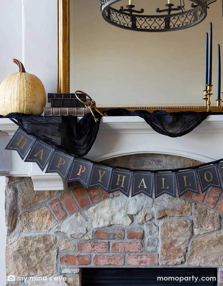 A close up shot of Momopartys's Happy Halloween banner hung over a Halloween mantel that's decorated with elegant Halloween decorations including a black and gold chandelier, black and gold candle holders, a beige colored pumpkin and a gold framed vintage mirror.