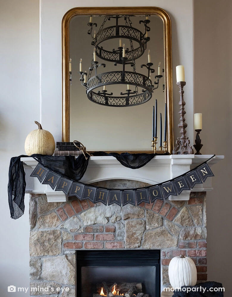 A Halloween fireplace mantel decorated with some elegant Halloween decorations including a black and gold chandelier, black and gold candle holders, some white and beige colored pumpkins and a gold framed vintage mirror. In the middle hung Momopartys's black and gold Happy Halloween banner by My Mind's Eye, making this a perfect inspo for a chic and elegant Halloween home decor.
