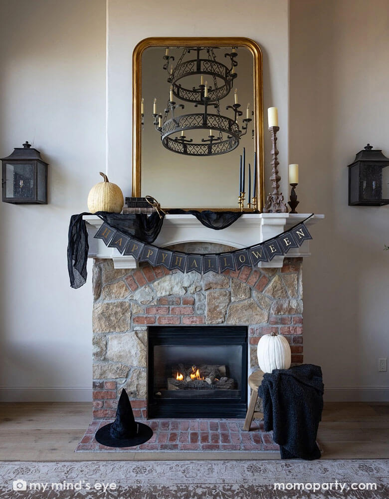A Halloween fireplace mantel decorated with some elegant Halloween decorations including a black and gold chandelier, black and gold candle holders, some white and beige colored pumpkins, a witch hat and a gold framed vintage mirror. In the middle hung Momopartys's black and gold Happy Halloween banner by My Mind's Eye, making this a perfect inspo for a chic and elegant Halloween home decor.