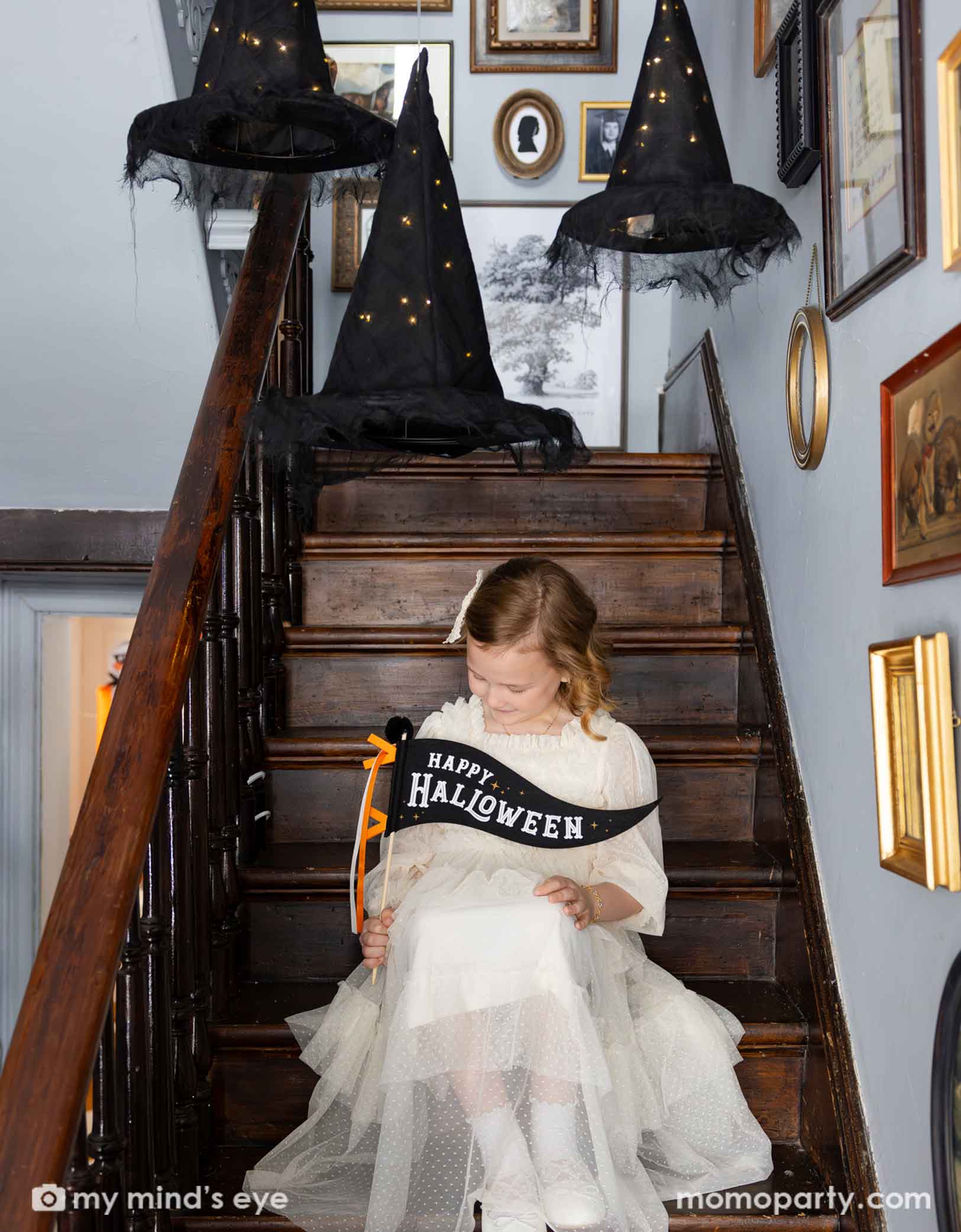 A girl in a vintage Halloween inspired white tulle dress sitting on a staircase holding Momopartys's 14" Happy Halloween felt party pennant looking down. Above her are three lit-up witch's hats hung from the ceiling. On the walls there are lots a vintage photo frame with old pictures, giving it a spooky vibe to a Halloween celebration.