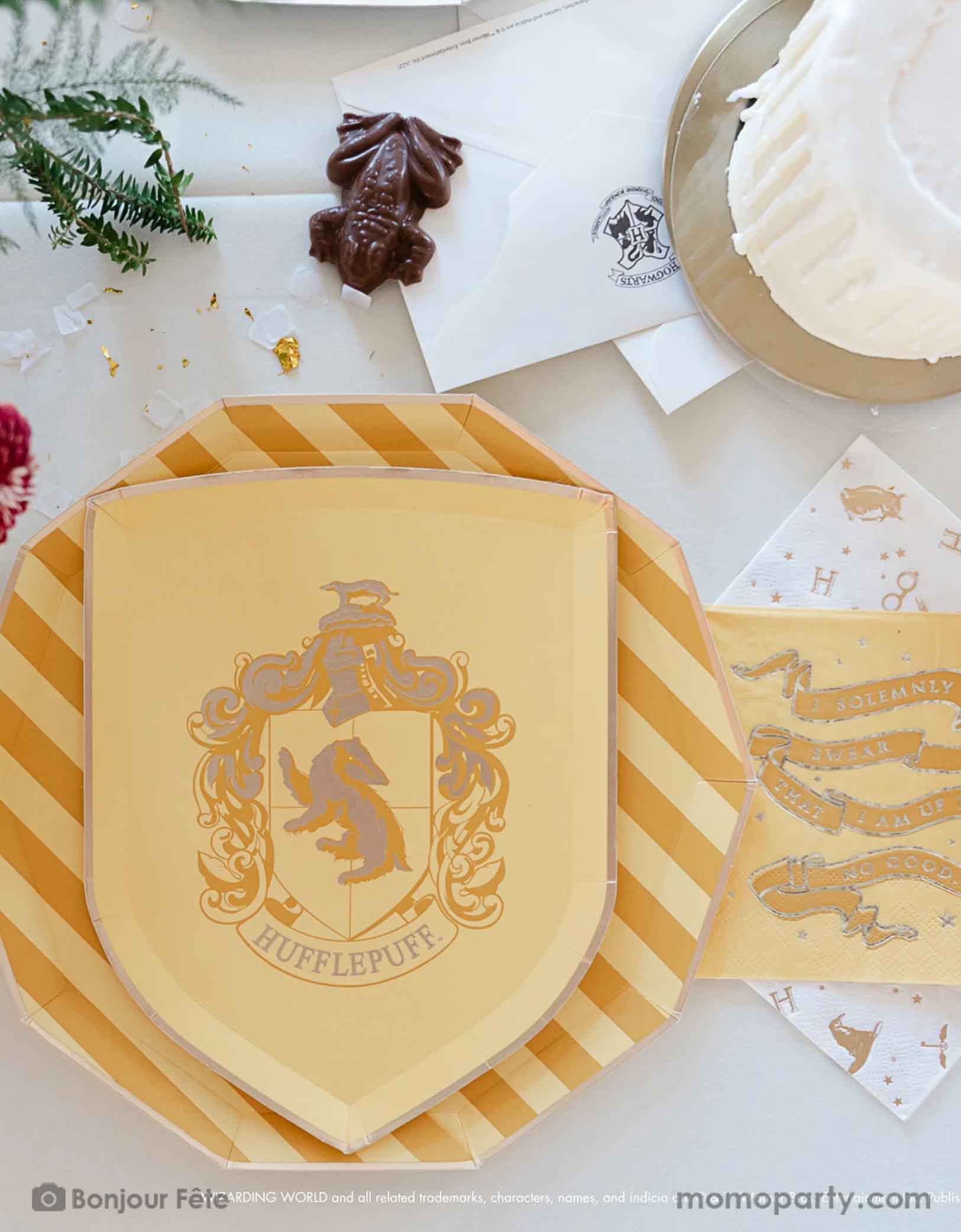 A beautiful and modern Harry Potter themed party tablescape features Momopartys's Harry Potter house pride shield-shaped 7" side plate with Hufflepuff logo and color paired with 10.25 green striped dinner plate. Next to the plates are Harry Potter motif large napkins in gold foil and Slytherin green small napkins. On the table are some letters from Hogwarts, a chocolate frog and a dripping candle birthday cake - making this a great inspo for a modern Harry Potter themed birthday party celebration.