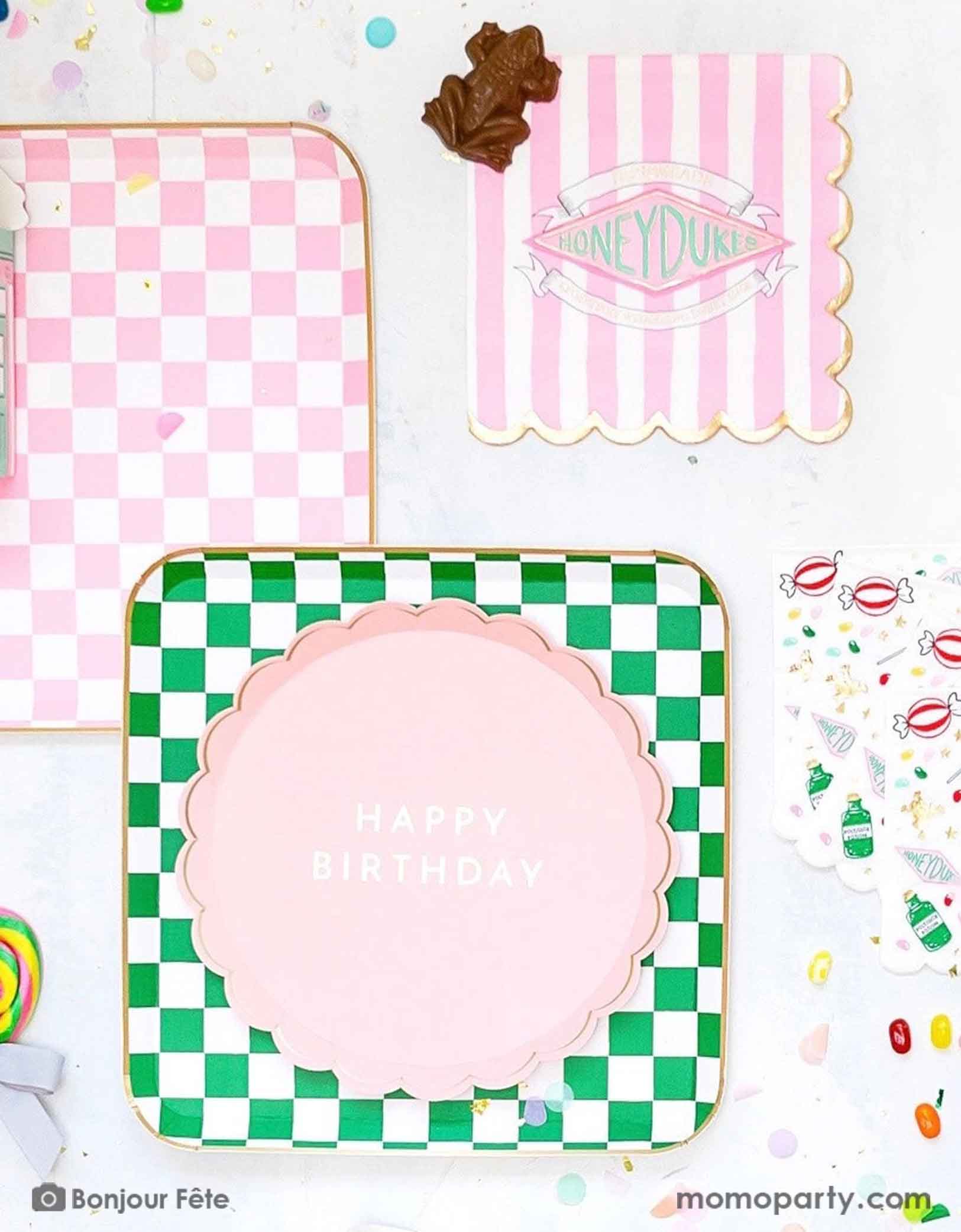 A Harry Potter themed tablescape featuring Momopartys's Harry Potter party collection including pink and green checkered dinner plates with pink Happy Birthday scalloped plate, next to them is pink striped Honeyduke large napkins with a chocolate frog. Along with some confetti and lollipops - a festive scene for kid's Harry Potter themed birthday celebration.