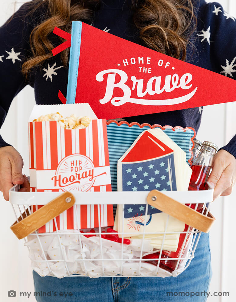 A lady holding an occasion bin filled with festive patriotic themed party goods including Momopartys's 15" Home of the Brave red felt pennant by My Mind's Eye, blue stripe scallop-edged plates, rocket shaped guest napkins, red striped popcorn treat boxes with "hip hip hooray" on it, these are perfect decorations for a Memorial Day celebration or 4th of July Independence Day party!