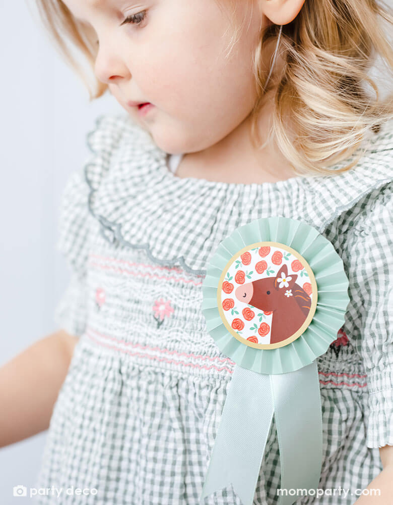 A close up shot of a preschool girl with a birthday party hat in a gingham dress and Momopartys's pastel mint floral horse rosette ribbon on her chest, a perfect decoration for girl's pony or horse themed birthday celebration.