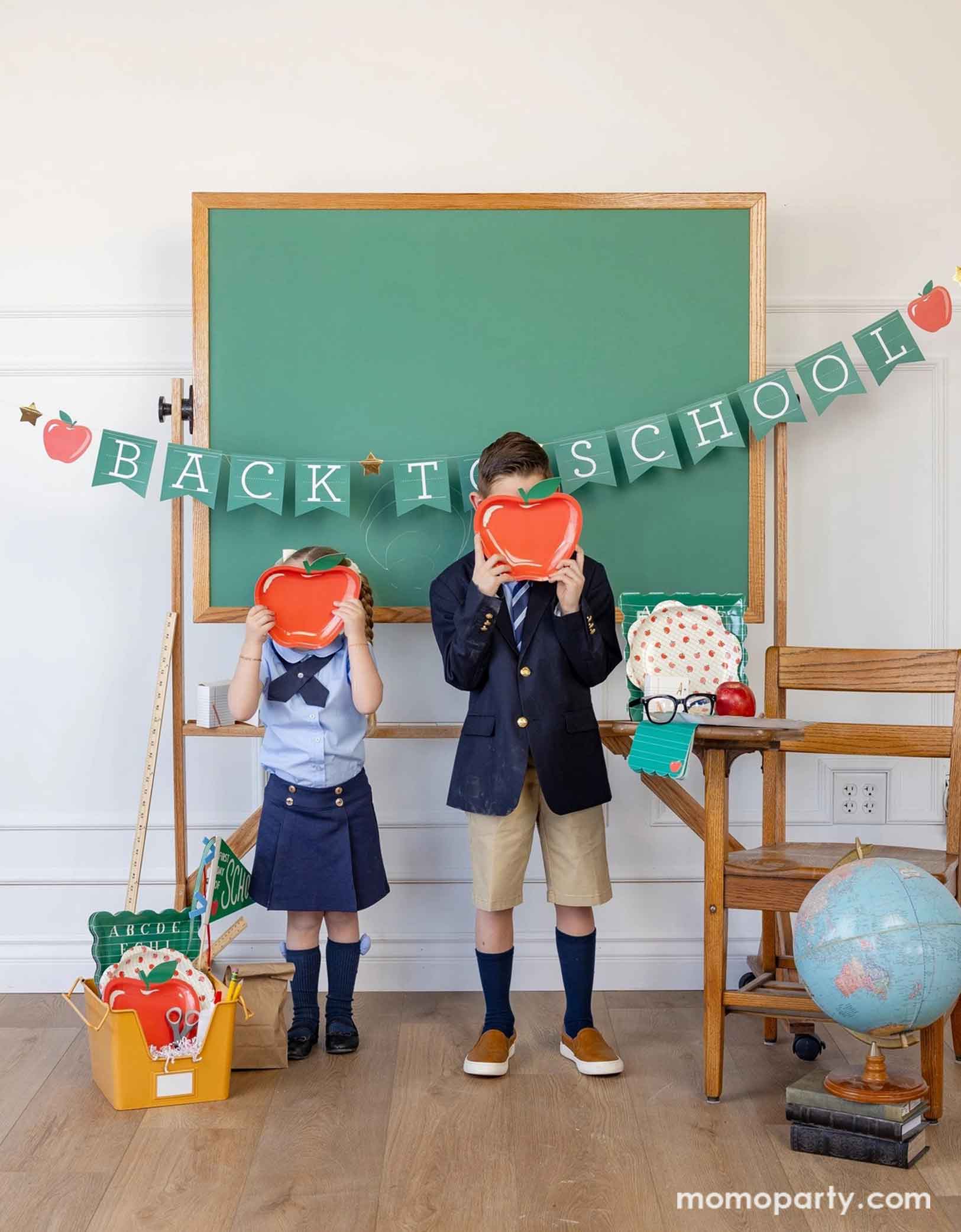 Two school-aged kids in their uniforms holding Momopartys's 8.5" apple shaped plates standing in a classroom setting with a school chair, books and globe, decorated with a bunch of school themed party supplies from Momopartys including apple shaped plates, apple pattern plates, alphabet dinner plates, notebook treat boxes and matching napkins. Behind her is a classroom chalkboard with the apple back to school party banner hung across - an adorable and cute set up to celebrate kid's back to school season!