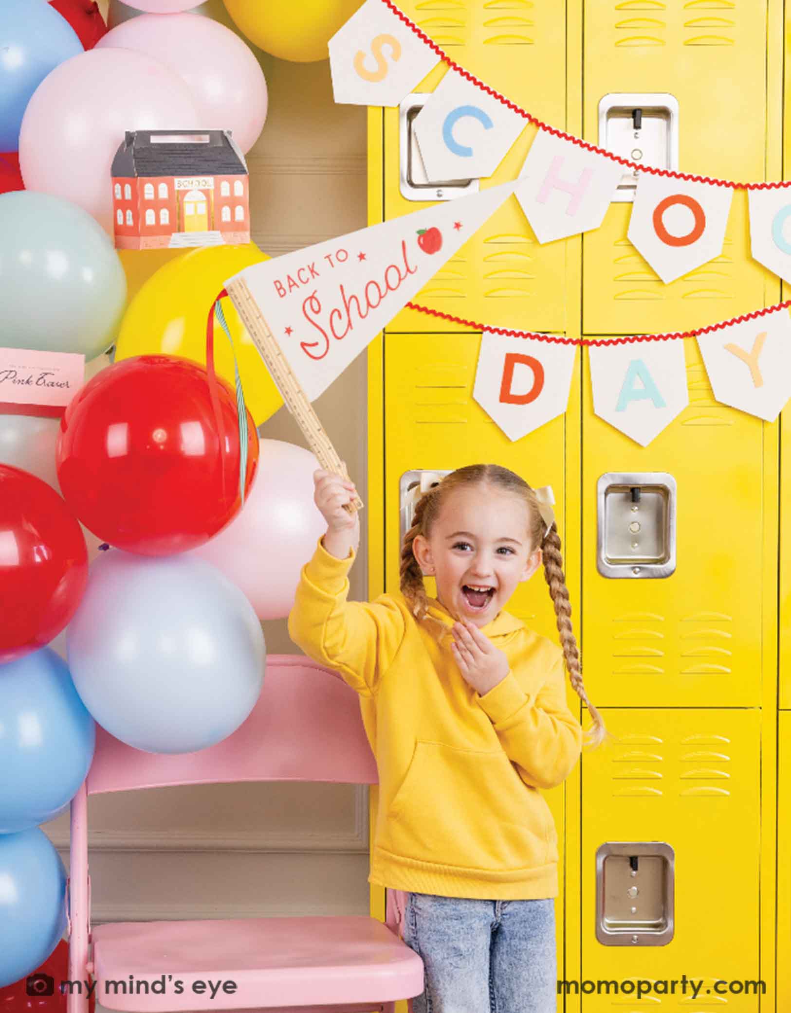 A girl in yellow shirt standing in front of a first day of school celebration decoration set up featuring various school themed party decors from Momopartys including School Days felt party banner and a festive balloon garland. She's holding the ruler back to school felt party pennant smiling happily. There's a yellow locker wall behind her and a pink folding chair. Making this a cute party decoration set up for kid's first day of school celebration.