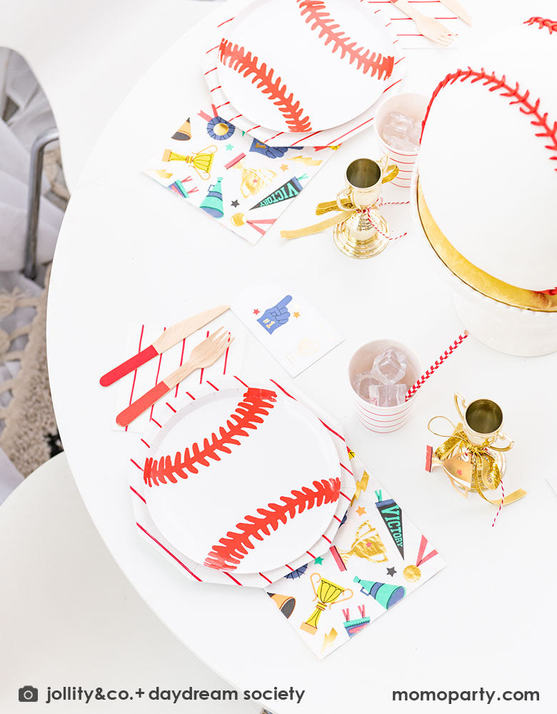 A sports themed party tablescape featuring Momopartys's 7.5 inch baseball plate layered on top of red striped plates, napkins and sports themed napkins by Daydream Society. In the middle of the table there's a baseball shaped cake which is surrounded by mini trophies, all for a fun sports champion celebration for kids!