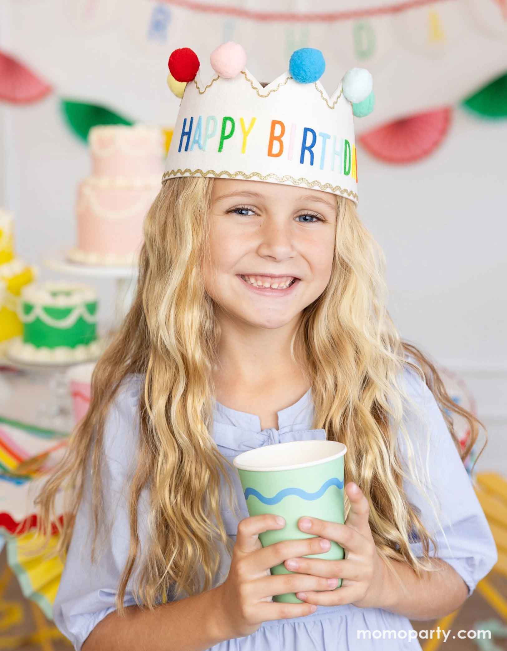 A school-aged girl wearing a colorful felt happy birthday crown with pom poms holding Momopartys's 12 oz green Ric Rac party cup in her hands. In the back there's a table full up birthday cakes in different pastel colors and some festive happy birthday banners on the wall, making this a charming scene for kid's birthday party celebration.