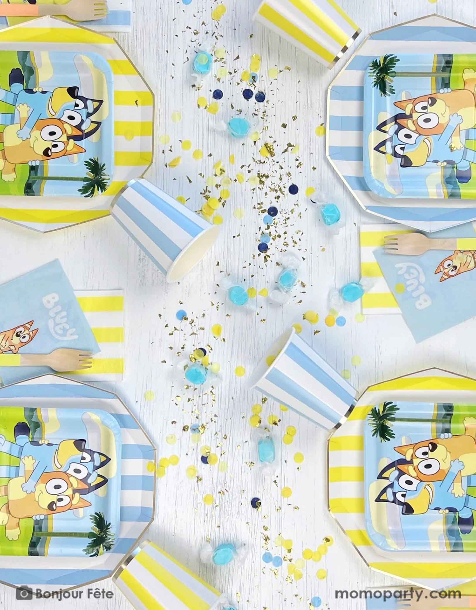 An overhead shot of a Bluey themed kid's birthday party table featuring Momopartys's modern blue and yellow cabana striped tablewares including paper plates, paper cups and napkins. On the dinner plates there are square side plates and party napkins featuring Bluey charcters including Bluey and Bingo, with matching colored confetti and blue colored candies on the table.