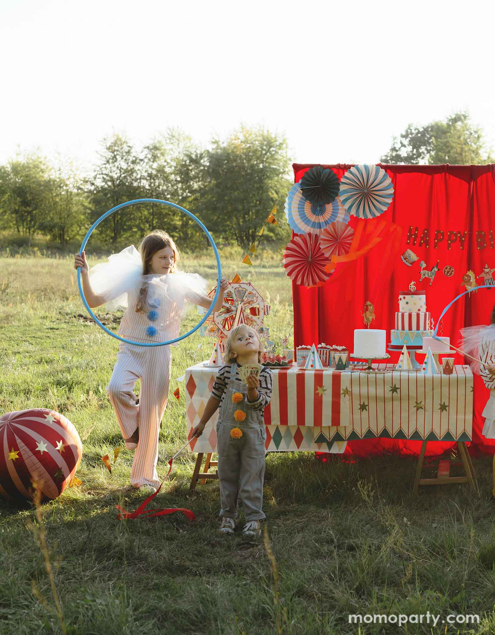 A vintage circus themed party set up in the park featuring various carnival circus themed party decorations from Momopartys including the red striped globe foil balloon next to the red curtain party backdrop. In the front there's a long table with goodies covered with circus themed table covers. A couple of kids dressed in vintage carnival themed clothing playing hula loops, making this a whimsical and playful setting for a fun kid's circus party.