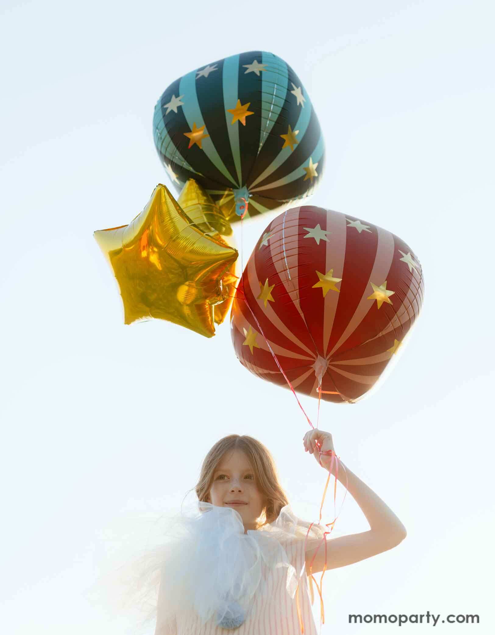 A girl dressed in quirky vintage circus inspired clothing holding Momopartys's circus themed party balloons. The foil balloons floating in the air include a red globe foil balloon, a green striped globe foil balloon and some gold star foil balloons, perfect decorations for a fun kid's circus/carnival themed celebration.