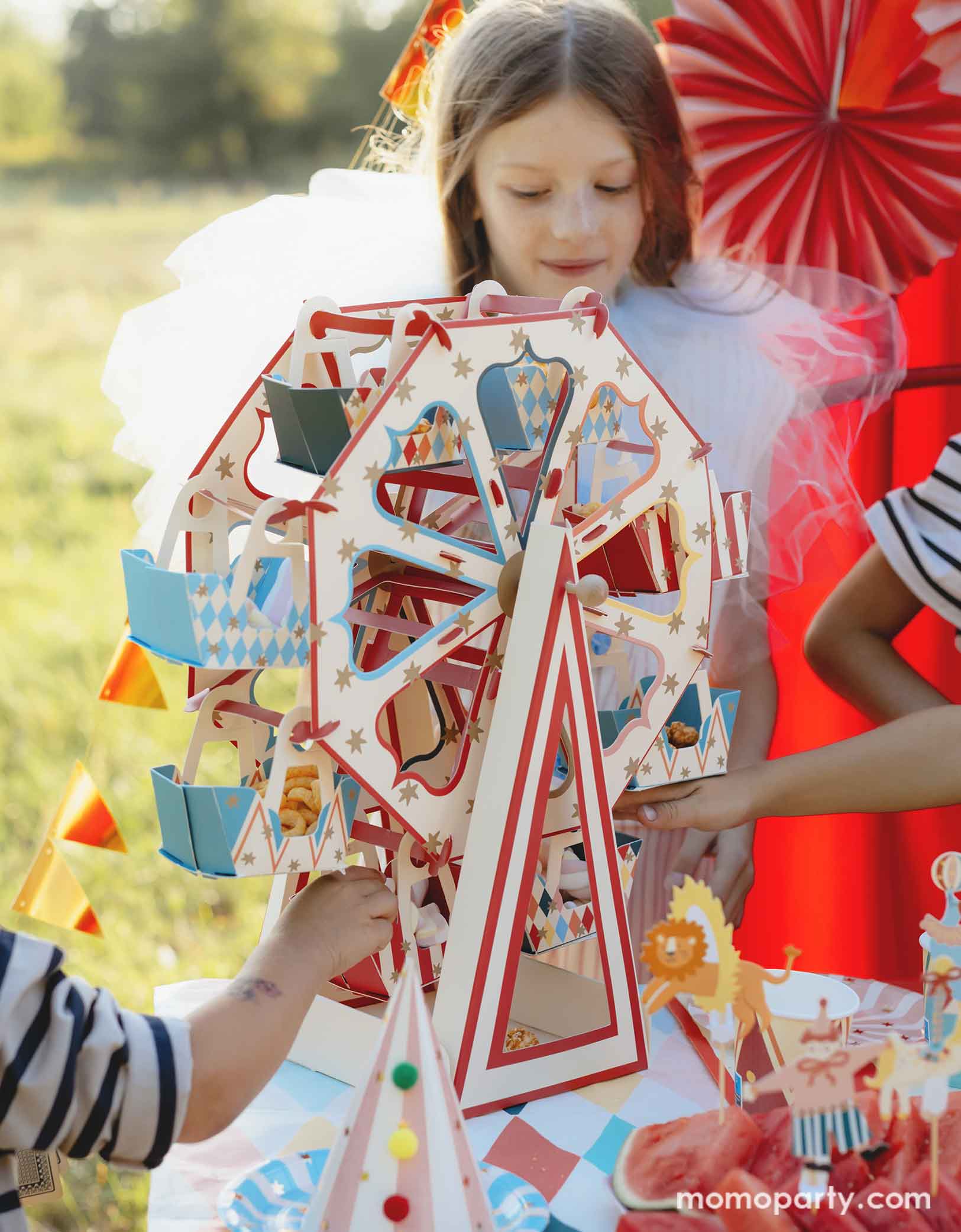 A few kids gathering at the party table in a circus & carnival themed birthday party which features Momopartys's 18.9" x 19.7" Carnival Ferris Wheel Snack Stand by Party Deco. This stunning centerpiece with its circus-inspired design is loaded with treats and cupcakes and they kids are grabbing the goodies from this fun treat stand. In the back, there's a red stage curtain as a party backdrop decorated with circus themed birthday banner with circus animal party pennants and colorful party fans.