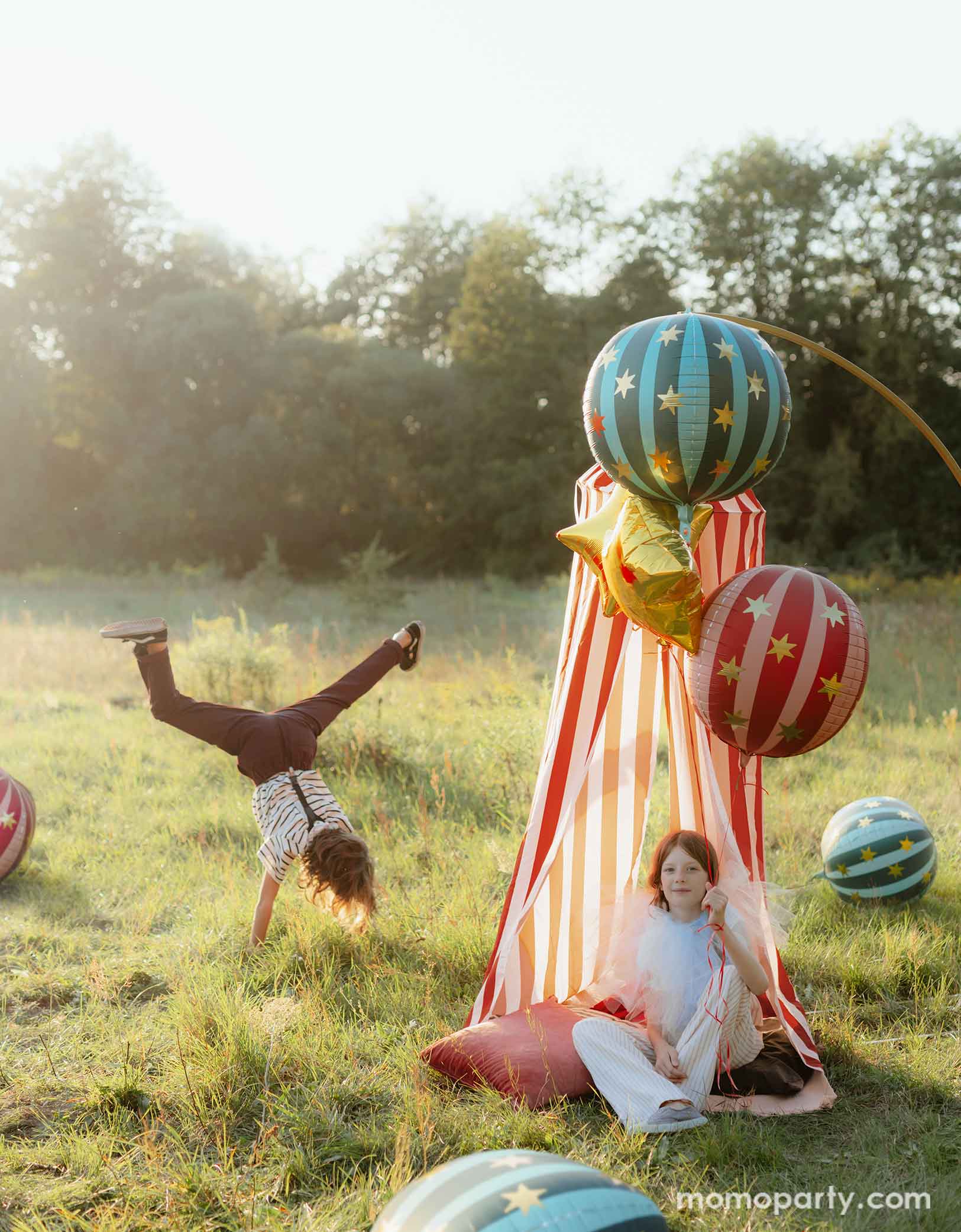 Two kids dressed in vintage and quirky clothing hanging out and playing in a vintage circus themed party featuring various carnival circus themed party decorations from Momopartys including the red striped globe foil balloon, the green striped globe foil balloon and gold star foil balloons floating next to a red and white striped circus themed play tent for kids. With sun dawn lighting from the back, making this a whimsical look for a fun circus/carnival themed celebration.