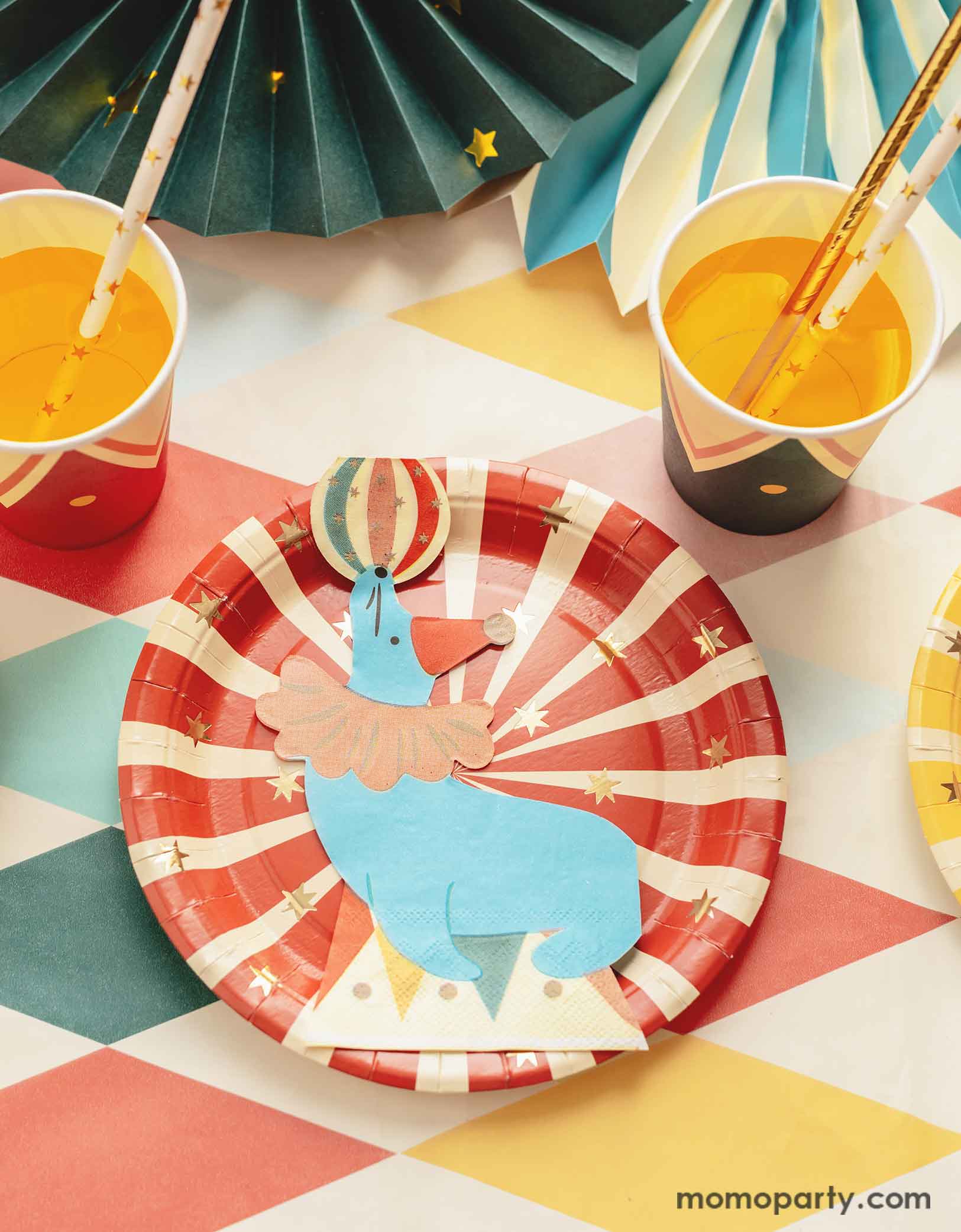 A carnival/circus themed part table featuring Momopartys's circus themed party tableware including a red striped paper plate and a sea lion playing with a ball shaped napkin, with matching party cups with gold foil star paper straws and some festive party fans on a circus themed striped table cover - making this great table setting idea for a fun and festive kid's carnival themed birthday celebration!