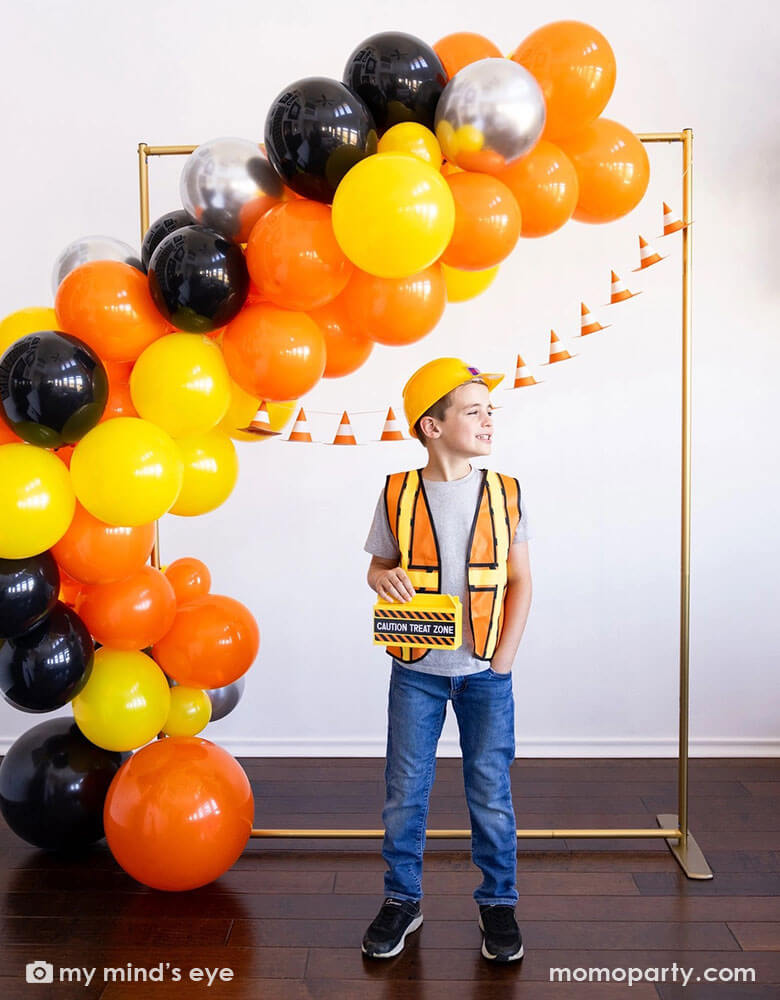 A school aged boy in his construction costume and hard hat holding Momopartys's toolbox shaped treat box by My Mind's Eye standing in front of a festive construction themed party decoration which features a balloon garland in black, orange silver and yellow and a construction cone banner hung over on the wall - a perfect decoration idea for kid's construction builder themed birthday celebration party.