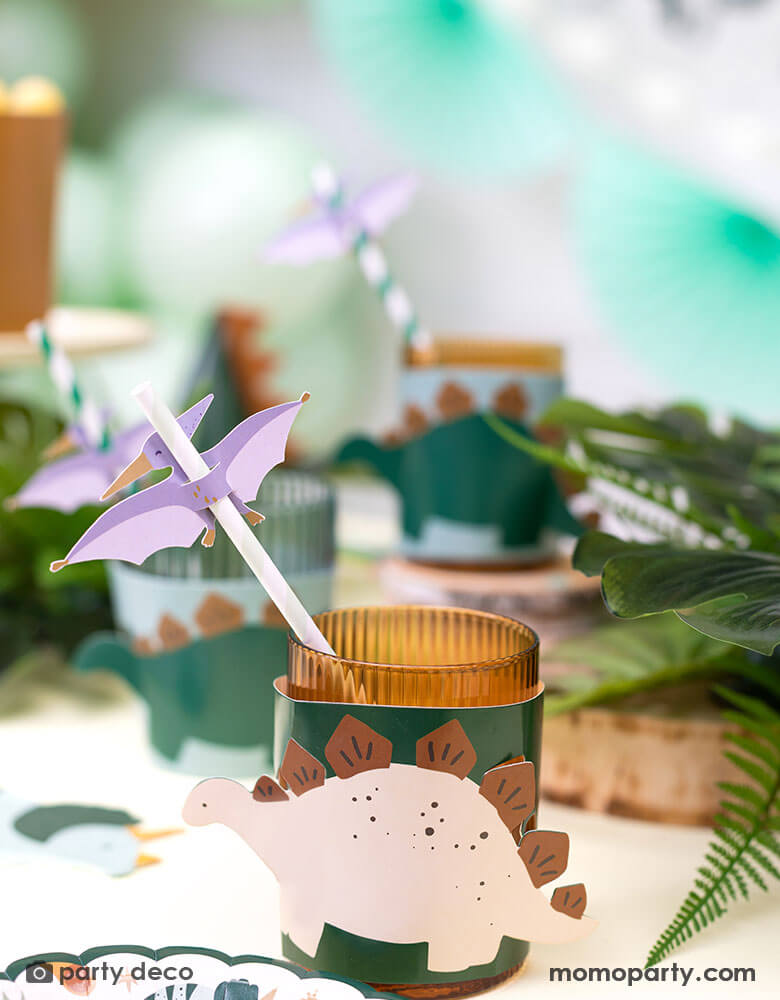 A dinosaur themed birthday party table featuring Momopartys's dinosaur cup sleeves and Pterodactyl paper straws, with tropical themed plants around the table, making this a great inspo for a modern and stylish kid's dinosaur themed party.