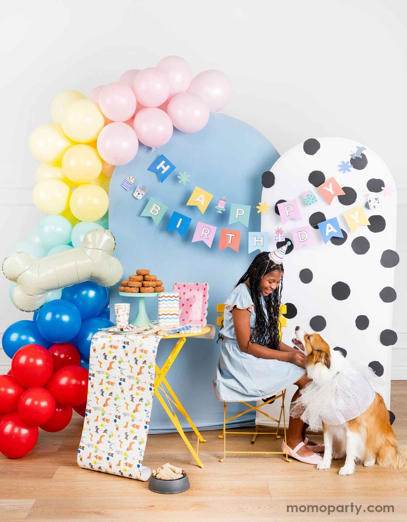 A dog themed party decoration set up featuring Momopartys's dog themed birthday supplies by My Mind's Eye. Including dog Happy Birthday party banner hung over a polka dotted and a pastel blue party backdrop with a pastel colored party balloon garland and a bone shaped foil balloon. On the table there are some dog themed party tablewares sugar cookies, treats and a donut tower. A girl in a birthday hat is sitting on the side and petting her dog happily celebrating her birthday.