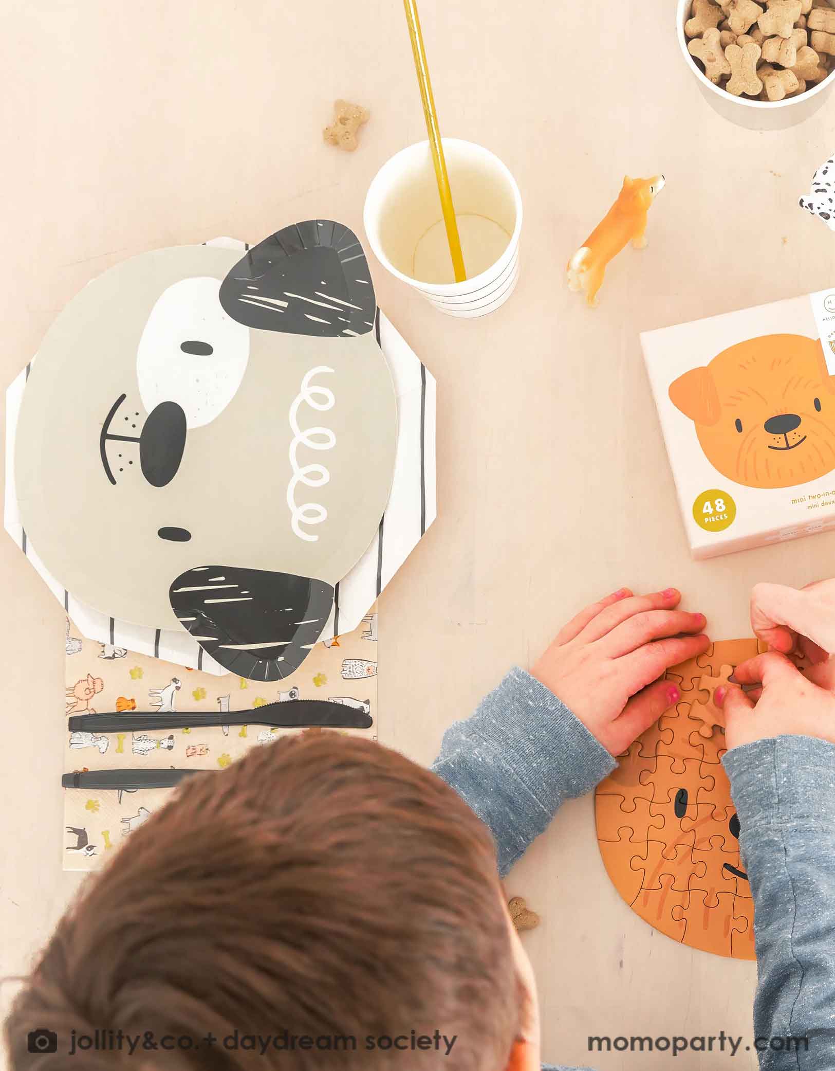 A little boy playing a puppy head shaped puzzle on a beige dining table featuring Momopartys's cute dogs head shaped paper plate paired with a black striped paper plate, along with a napkin in modern illustrations with a warm neutral color palette of dog illustrations, with black utensil set. These modern Dog Birthday Party Supplies are perfect for a dog lover, a dog themed birthday party, Let's Pawty birthday party, adorable puppy themed party.