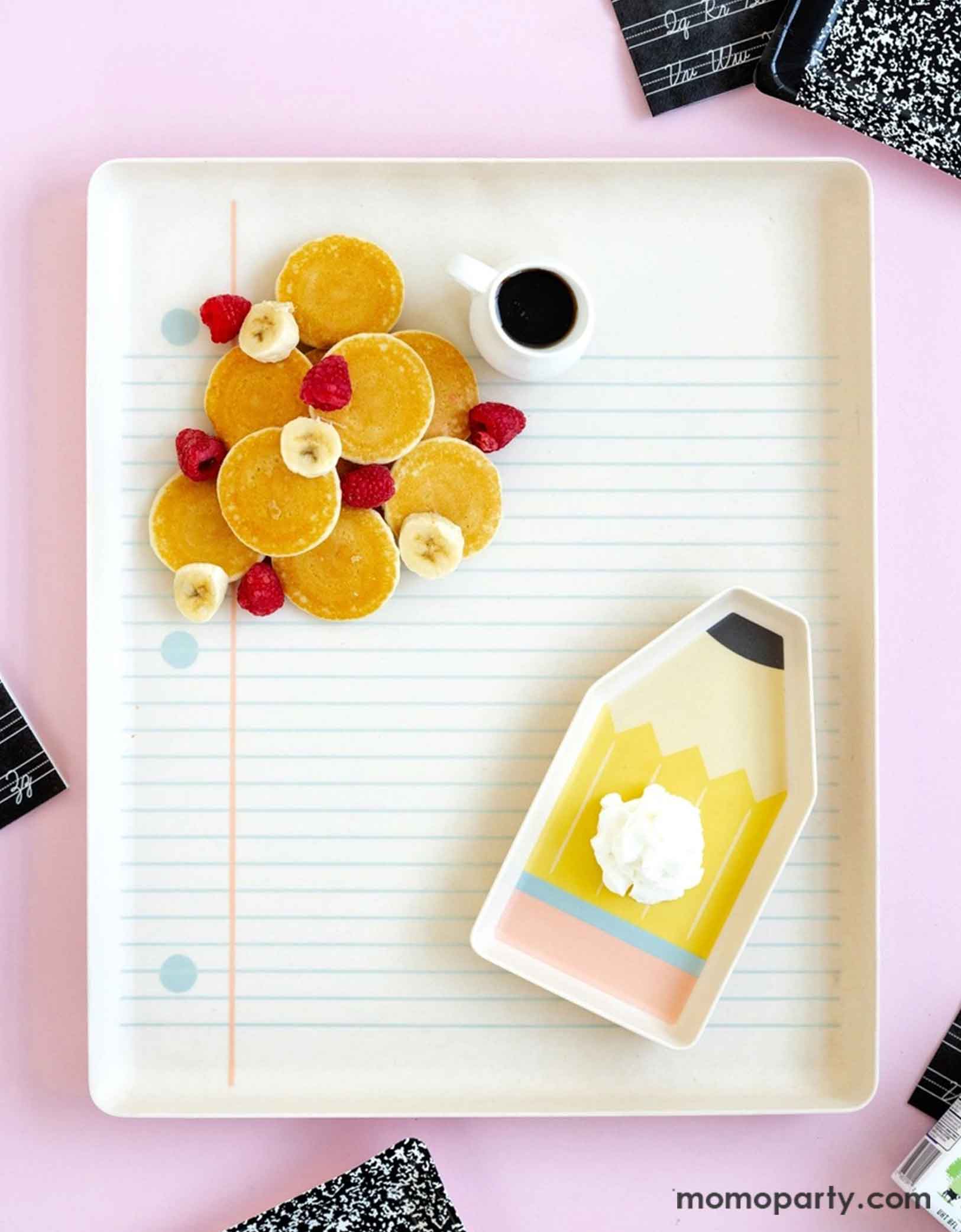 An overhead shot of Momopartys's 17" x 14" Paper and Pencil Bamboo Tray Set filled with a bunch of petite pancakes, banana bites, raspberries and a small syrup pourer. On the notebook bamboo tray is the pencil shaped bamboo try with a dollop of whip cream. This makes a great inspiration idea for a special first day of school breakfast for kids to celebrate the new school year!