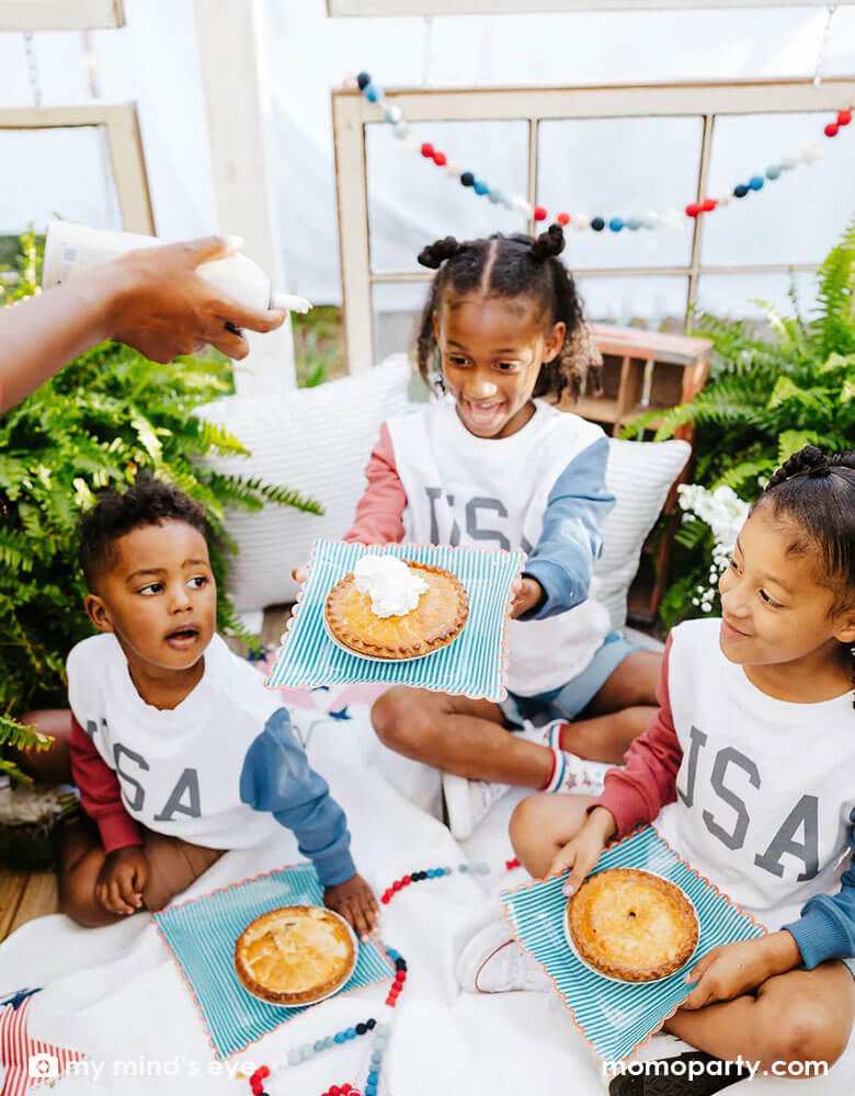 Three siblings in their USA sweatshirts holding Momopartys's red with blue stripe plates accented by a red scalloped edge. Designed by My Mind's Eye, these plates are in a perfect size for summer treats, including hand pies, hot dogs, burgers and more for a 4th of July cookout!