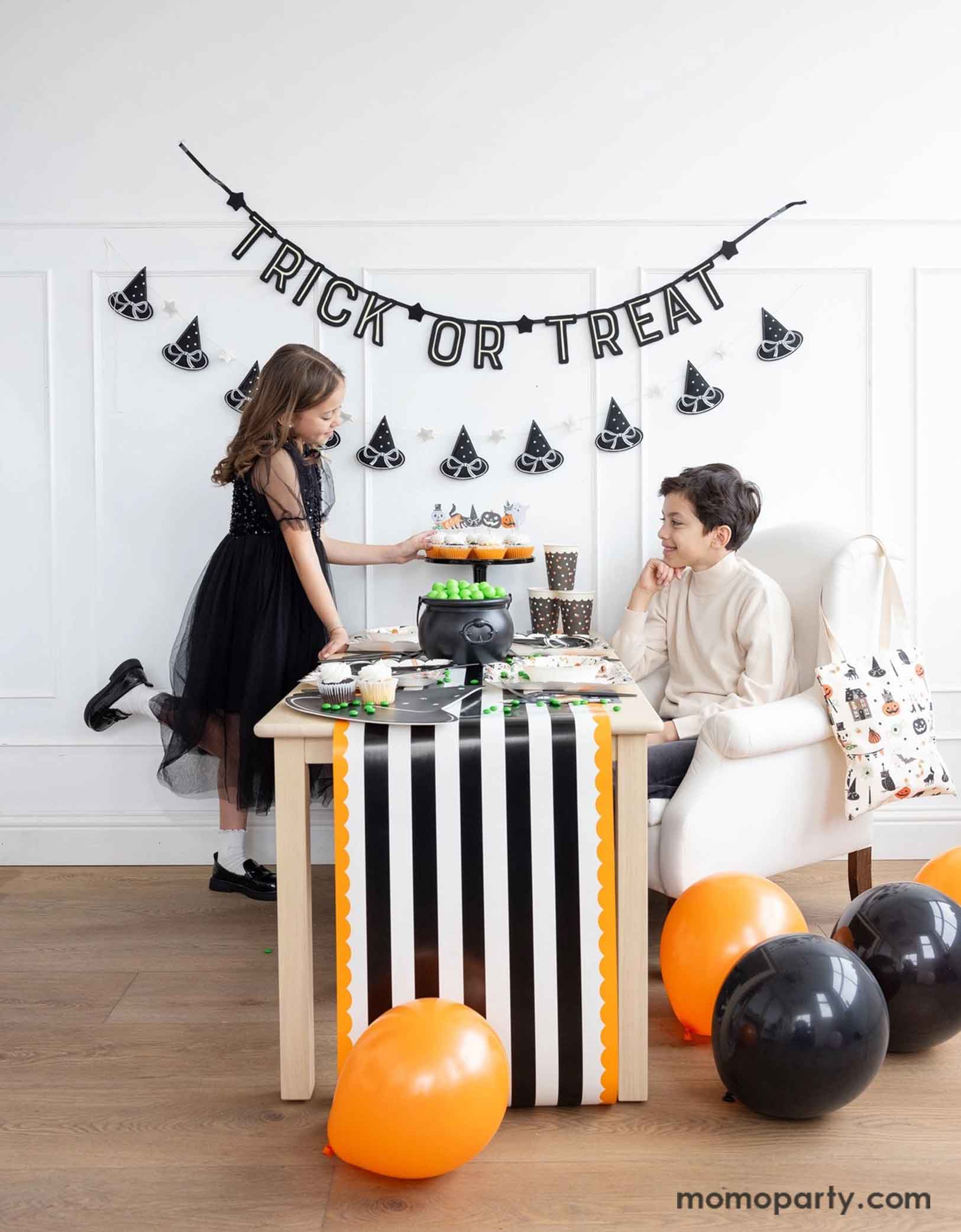 Two children at a Halloween-themed party with Momopartys's 'Trick or Treat' banner and the witch's hat garland hung on the white wall. There's a table decorated with multiple Halloween party supplies by Momopartys including witch's hat shaped bamboo tray, party cups and orange, black and white striped table runner draping from the table which some orange and black balloons next to. A cute and simple set up for kids to celebration Halloween at home.