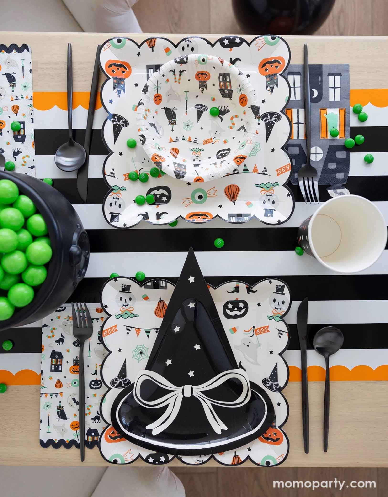 A overhead shot of a festive Halloween-themed table setting with the witch hat shaped plate, Trick or Treat dinner plates and bowls, the haunted mansion shaped napkins and black cutlery set on a black orange and white striped table runner fro Momopartys. On the table there's a cauldron filled with green gum balls and some green candies spread across the table. A perfect set up for kid's friendly Halloween celebration.