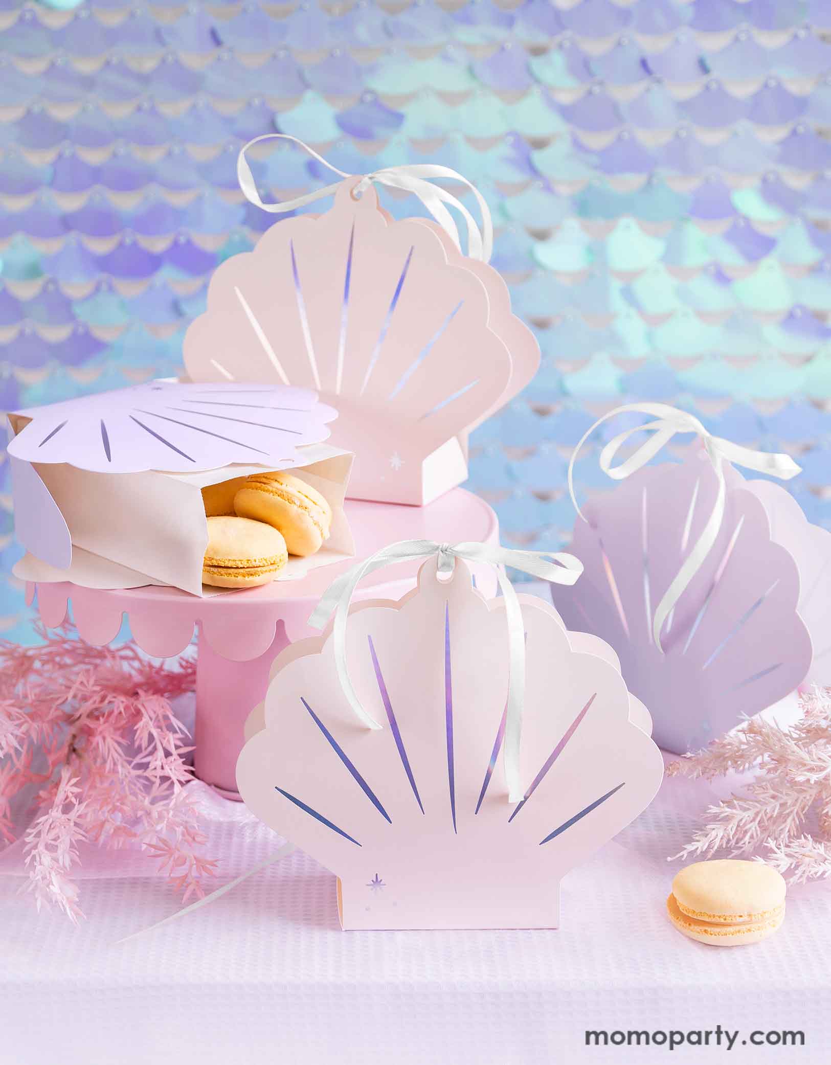 A mermaid birthday party table featuring Momopartys's 6.3" x 6.9" x 2.2" Seashell Treat Boxes by Party Deco. These boxes come in a set of 4 boxes in 2 colors of ombre pink and ombre lilac with a bow tied at the top. In the boxes there are some vanilla macarons on a pink cake stand surrounded by pink seagrass decorations. Behind the table there's a seashell-shaped iridescent sequin curtain as the backdrop, creating a dreamy and whimsical scene for a magical under the sea bash.